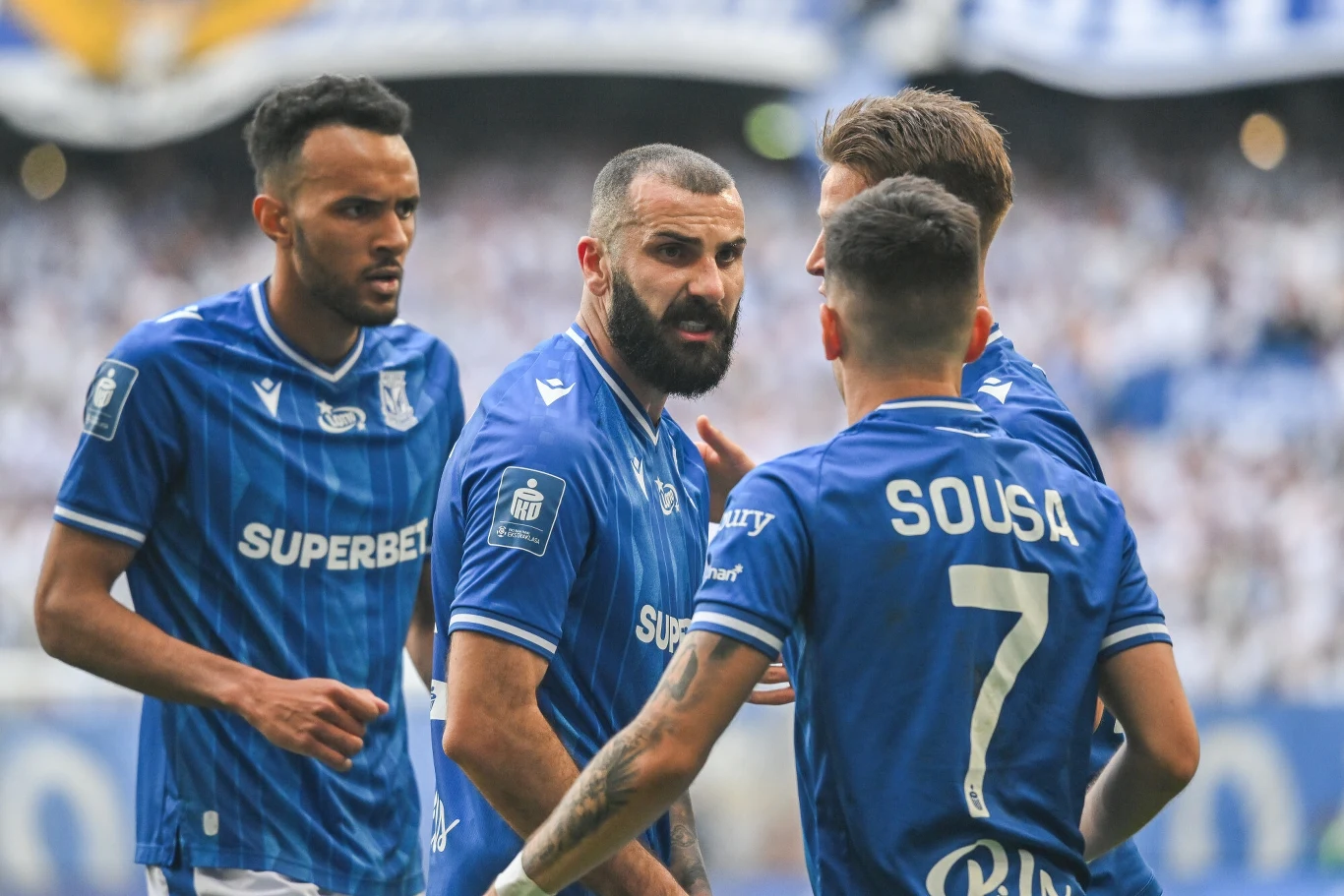 Lech Poznań Grupa piłkarzy w niebieskich koszulkach drużyny Lech Poznań podczas intensywnej wymiany zdań na boisku, widoczne skoncentrowane i emocjonalne twarze zawodników, tłum kibiców zamazany w tle.