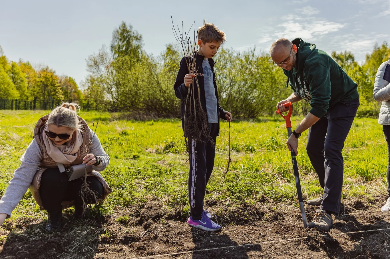 Fot. Miasto Gdańsk Grupa osób podczas sadzenia młodych drzewek na otwartym terenie, mężczyzna z łopatą kopie dół, chłopiec trzyma sadzonki, kobieta przykuca i pracuje przy ziemi, w tle zielone krzewy i drzewa.