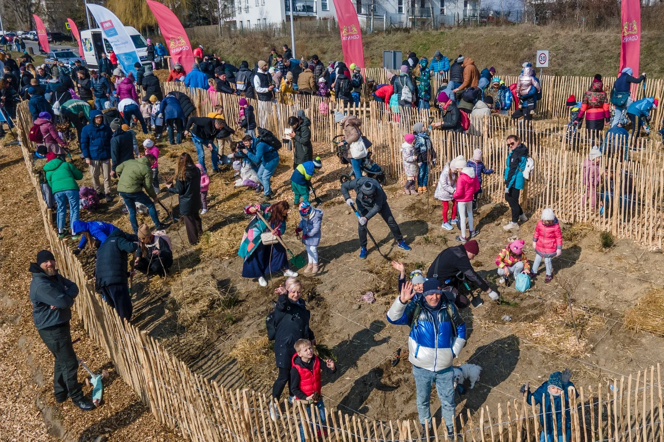 Fot. Grzegorz Mehring/ gdansk.pl Duża grupa ludzi, w tym rodziny z dziećmi, bierze udział w akcji sadzenia drzew na ogrodzonym terenie, wokół rozmieszczone są banery i ogrodzenie z jasnego drewna, atmosfera jest radosna i społeczna.