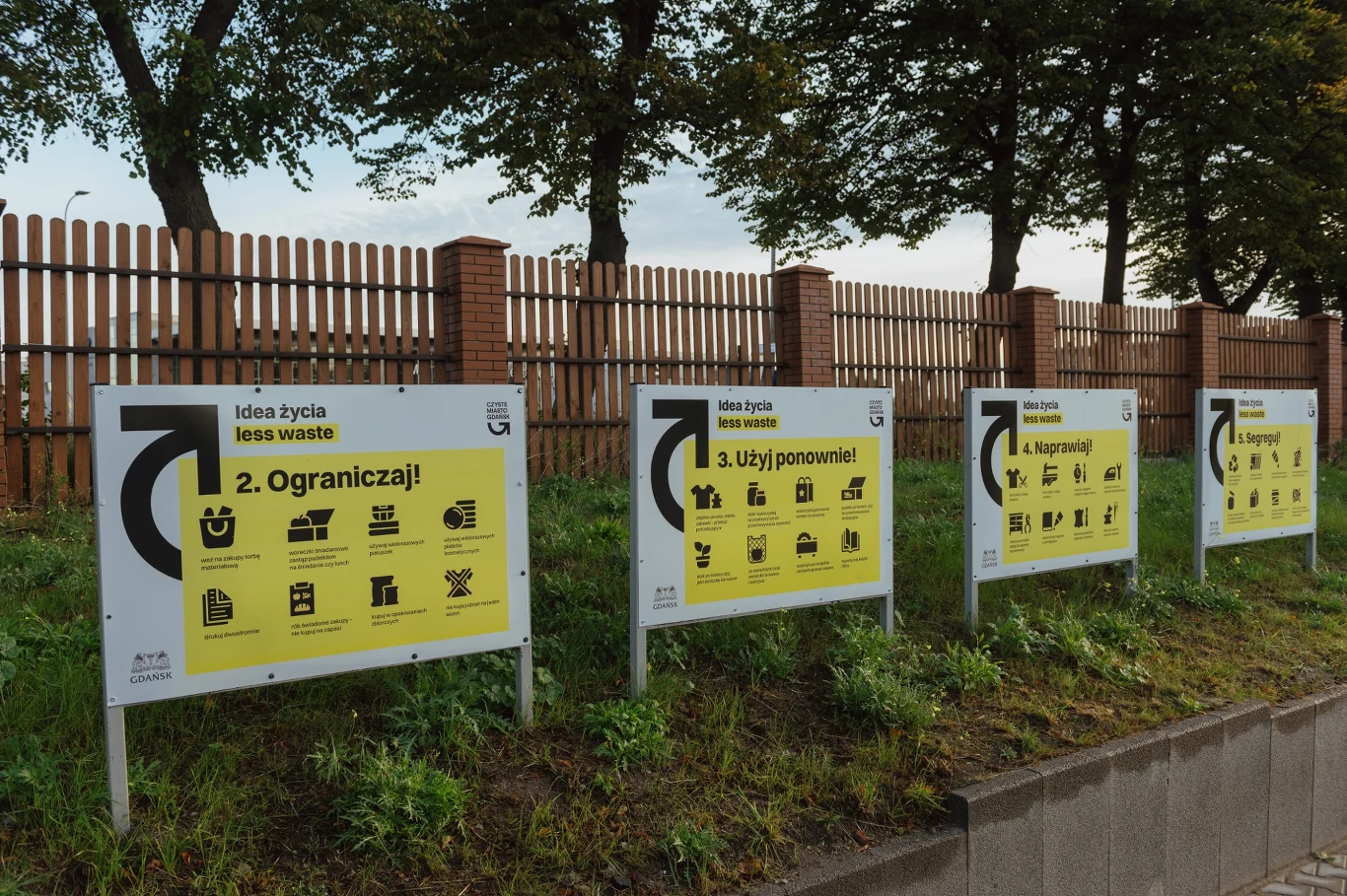 Edukacja ekologiczna w Gdańsku Cztery żółte tablice informacyjne z czarnym napisem i ikonami prezentujące zasady less waste ustawione przy ogrodzeniu z drewnianych sztachet i muru, z tyłu drzewa oraz trawniki.