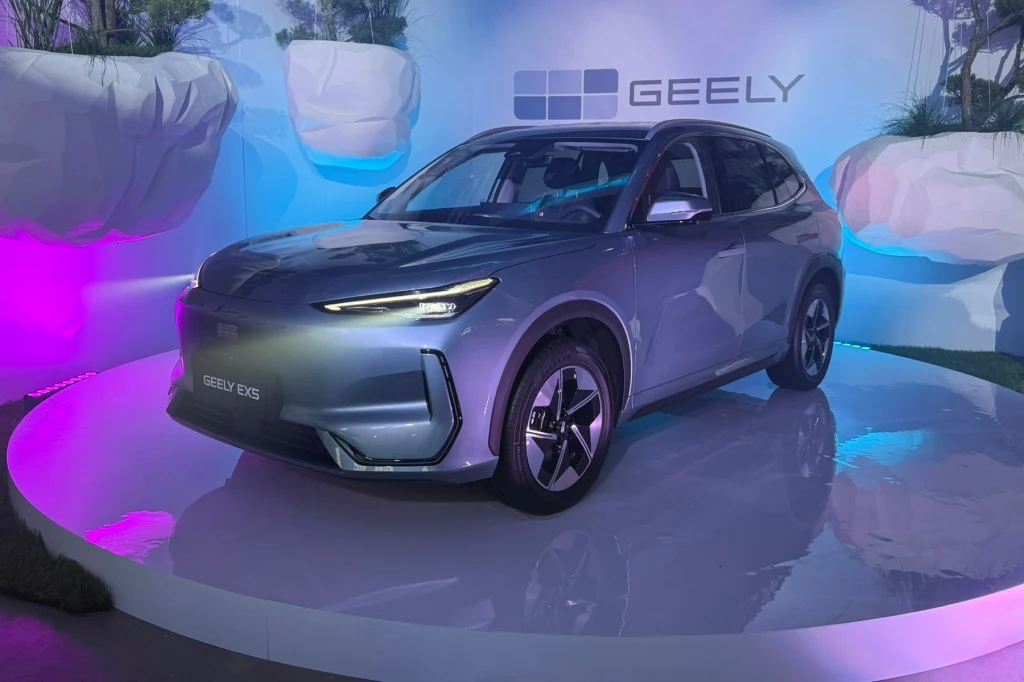 Koncern Geely przedstawia ambitne plany na nadchodzące lata. Nowoczesny srebrny samochód elektryczny Geely EX5 prezentowany na podświetlonym podium, w otoczeniu futurystycznej scenografii i kolorowego oświetlenia, z widocznym logotypem marki w tle.
