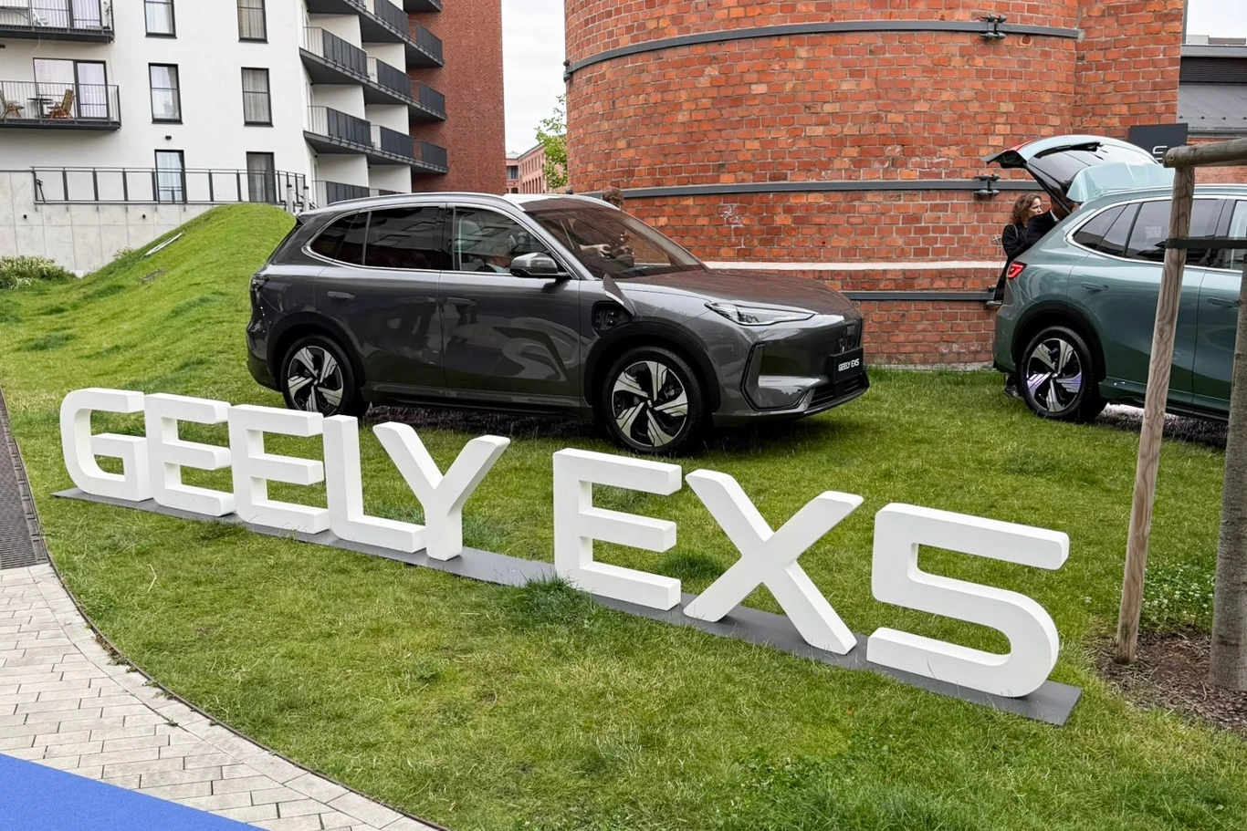 Nowoczesny samochód SUV marki Geely EX5 ustawiony na trawie obok dużego napisu z nazwą modelu, w tle widoczne są fragmenty budynków mieszkalnych oraz elementy miejskiej architektury.