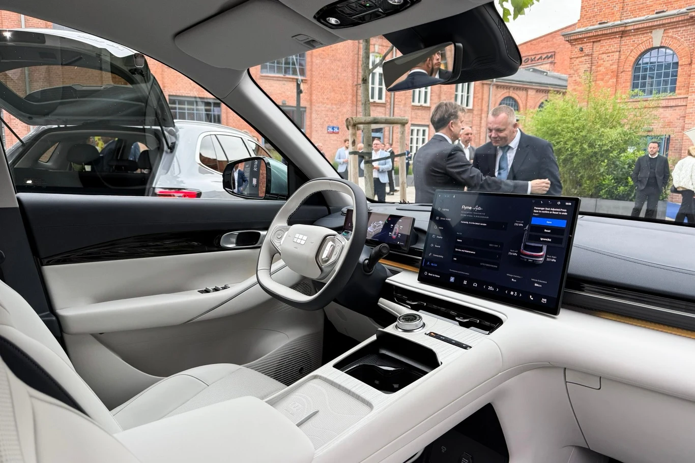 Nowoczesne wnętrze samochodu elektrycznego z jasną skórzaną tapicerką, dużym dotykowym wyświetlaczem centralnym oraz eleganckim wykończeniem kokpitu, na zewnątrz widać kilku eleganckich mężczyzn rozmawiających obok budynku z czerwonej cegły.