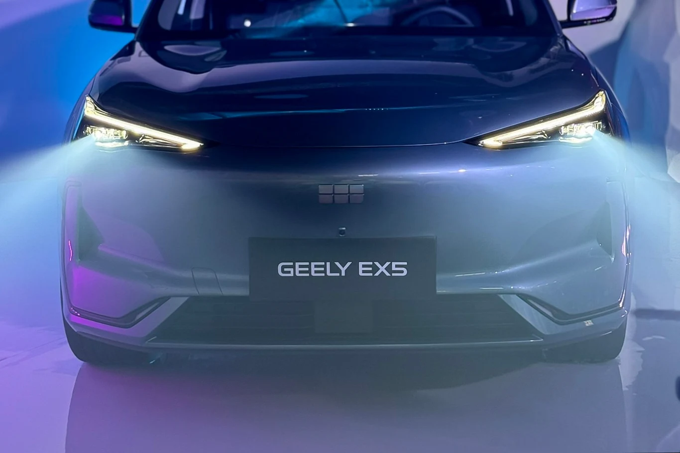 Przód nowoczesnego samochodu Geely EX5 z włączonymi światłami mijania, podkreślony niebieskim oświetleniem oraz refleksami na karoserii.