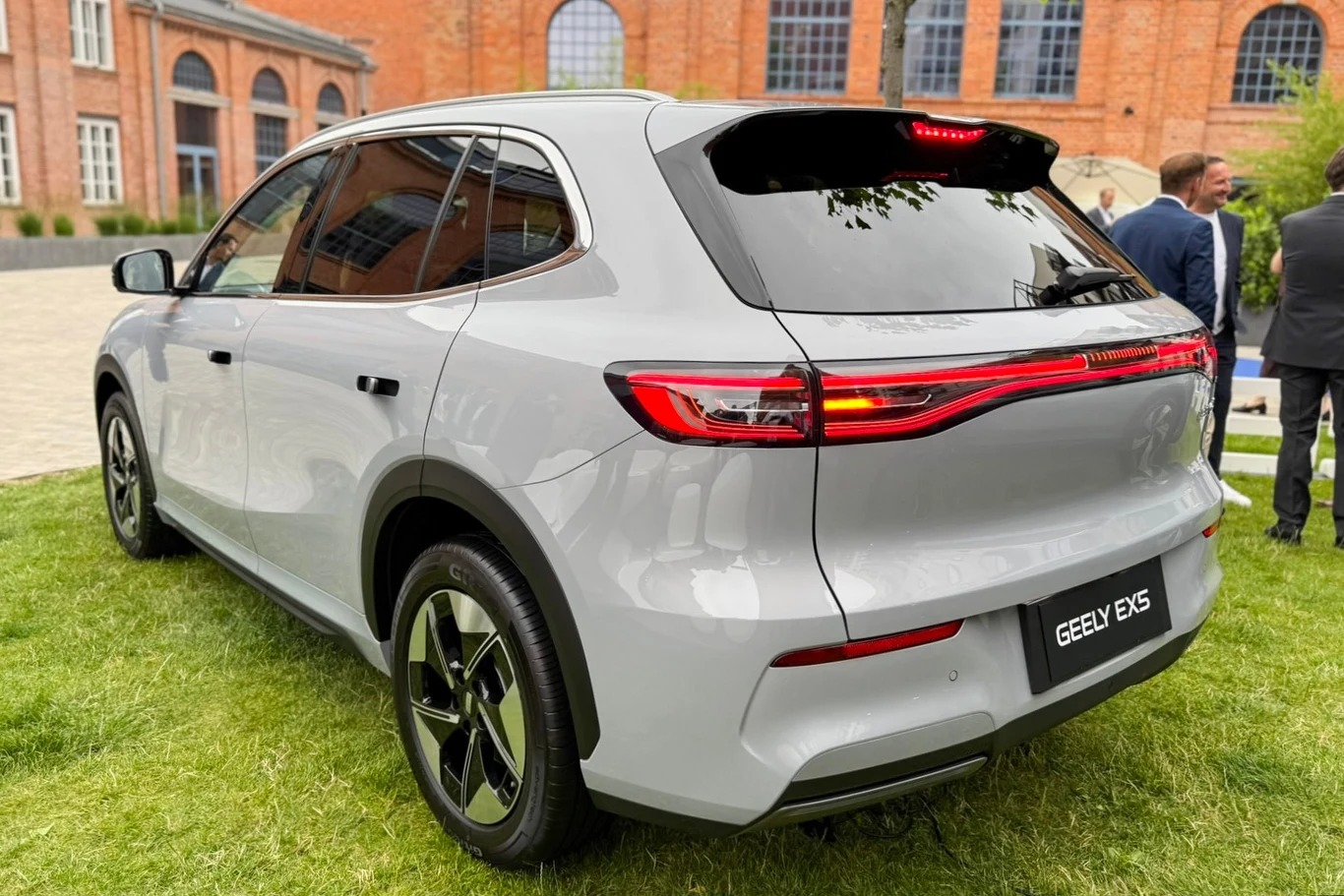 Srebrny samochód elektryczny marki Geely EX5 zaparkowany na zielonej trawie, w tle budynki z czerwonej cegły oraz kilka osób stojących na chodniku.