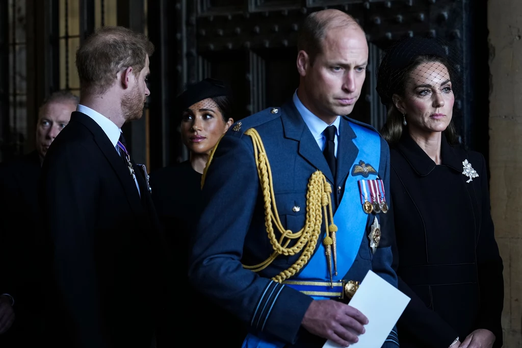 Sytuacja między Harrym a resztą członków rodziny królewskiej jest napięta od dawna Grupa elegancko ubranych osób pogrążona w poważnym nastroju, mężczyzna w mundurze z odznaczeniami, obok kobiety w czarnych strojach żałobnych, pośród nich atmosfera skupienia i dostojności.