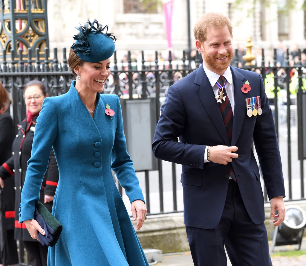 Niegdyś Harry miał doskonały kontakt chociażby ze swoją bratową, księżną Kate Uśmiechnięta kobieta w eleganckim niebieskim płaszczu i kapeluszu oraz mężczyzna w garniturze z medalami przypiętymi do marynarki spacerują razem na tle ogrodzenia i budynku w stylu klasycznym.