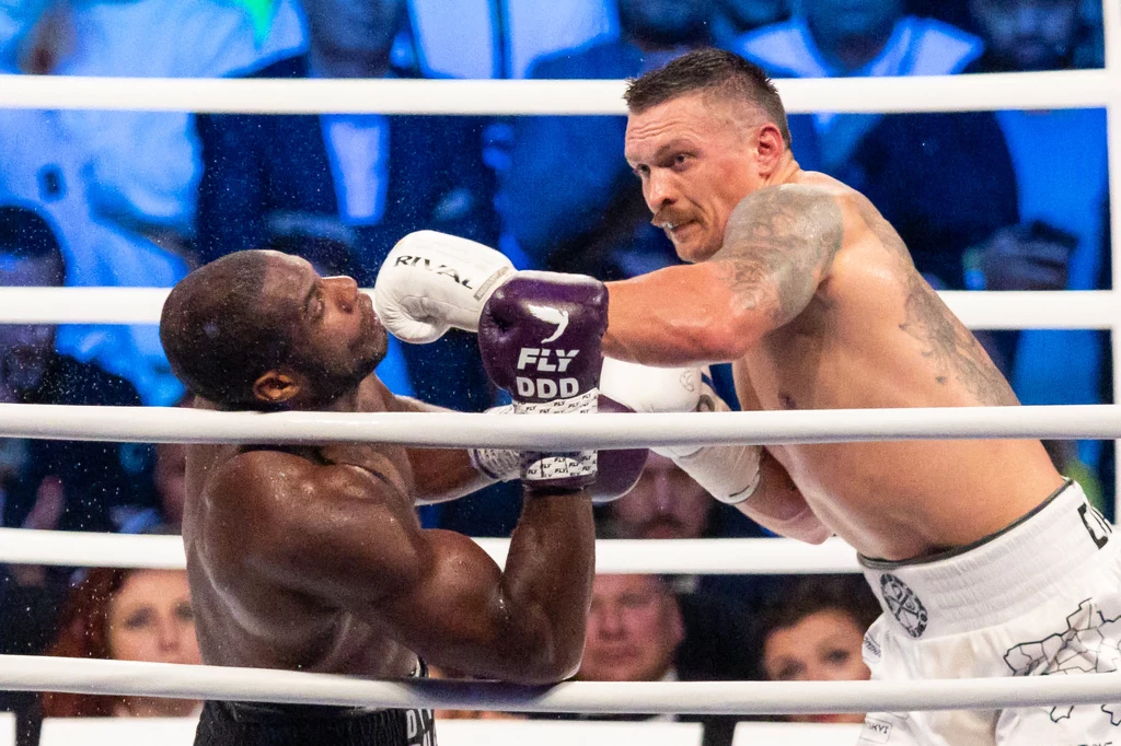 Dwóch bokserów podczas intensywnej wymiany ciosów w ringu, jeden z zawodników trafia drugiego mocnym ciosem w twarz, widoczne napięcie i dynamika sportowej rywalizacji, w tle publiczność na trybunach.