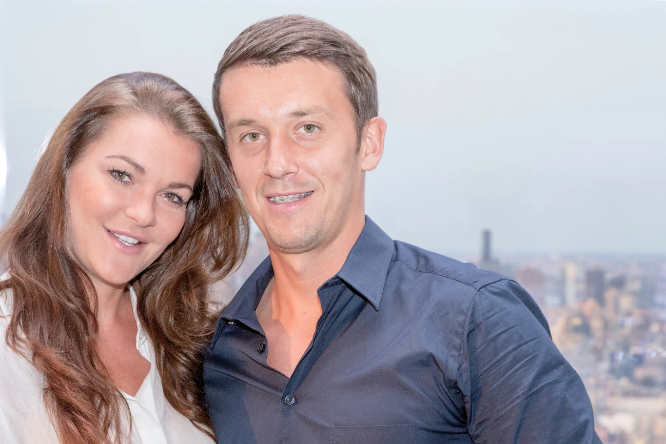 Agnieszka Radwańska i Dawid Celt Dwie uśmiechnięte osoby, kobieta o długich brązowych włosach oraz mężczyzna w niebieskiej koszuli, stoją blisko siebie na tle panoramy miasta.
