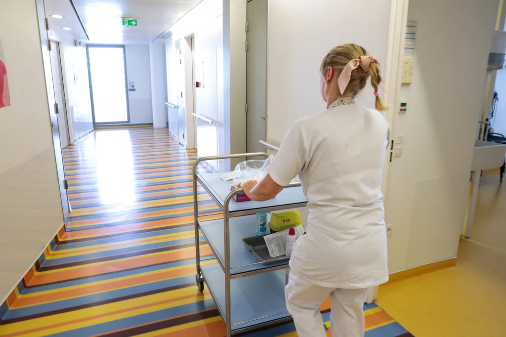 Korytarz szpitalny z pielęgniarką ubraną na biało, prowadzącą wózek z materiałami medycznymi; podłoga kolorowa w pasy, wnętrze jasne, schludne i uporządkowane.