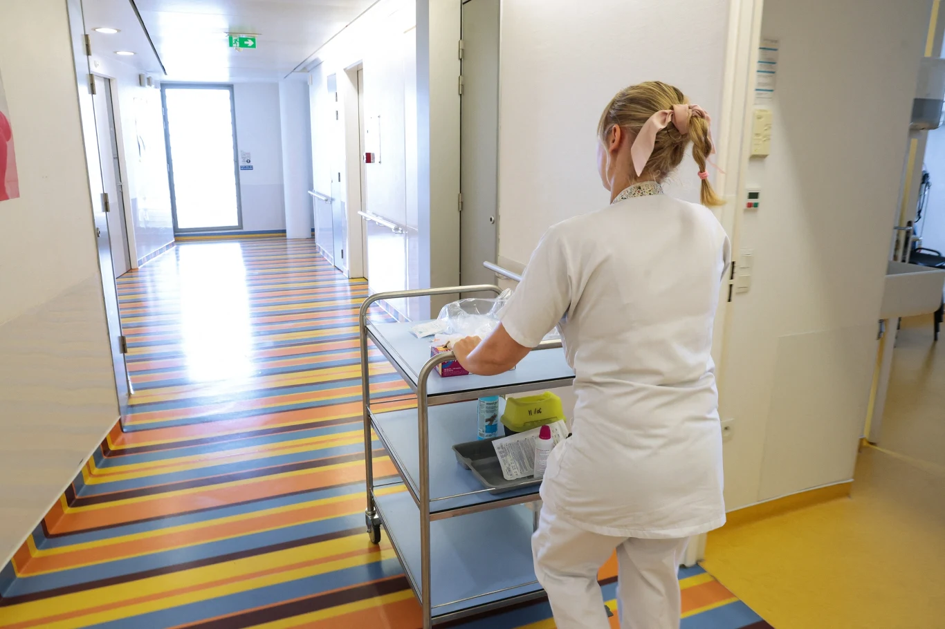 W pracy pielęgniarek zazwyczaj potrzebny jest średni poziom bezpieczeństwa psychologicznego Korytarz szpitalny z pielęgniarką ubraną na biało, prowadzącą wózek z materiałami medycznymi; podłoga kolorowa w pasy, wnętrze jasne, schludne i uporządkowane.