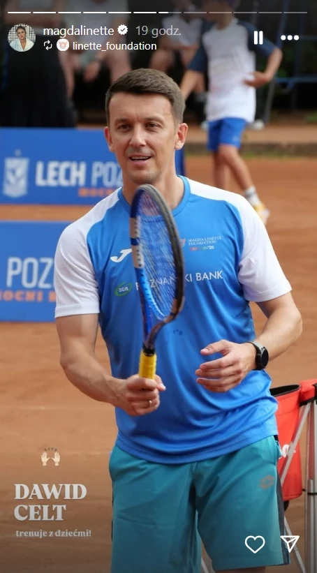 Dawid Celt na wydarzeniu fundacji Magdy Linette Mężczyzna ubrany w sportowy strój stoi na korcie tenisowym, trzymając rakietę do tenisa i uśmiechając się, w tle niewyraźne postaci oraz tablice sponsorskie.