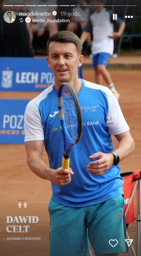Dawid Celt na wydarzeniu fundacji Magdy Linette Mężczyzna ubrany w sportowy strój stoi na korcie tenisowym, trzymając rakietę do tenisa i uśmiechając się, w tle niewyraźne postaci oraz tablice sponsorskie.
