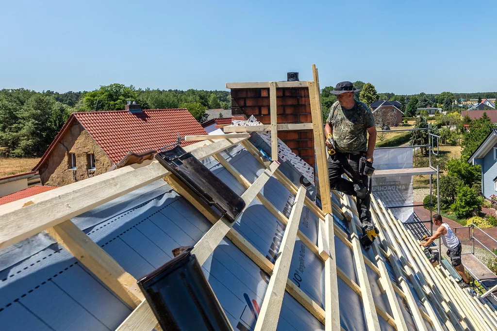 Dwóch pracowników wykonuje prace dekarskie na stromym dachu budynku jednorodzinnego, w tle widoczna wiejska zabudowa i drzewa, na pierwszym planie konstrukcja drewnianego szkieletu dachu z narzędziami oraz materiałami budowlanymi.