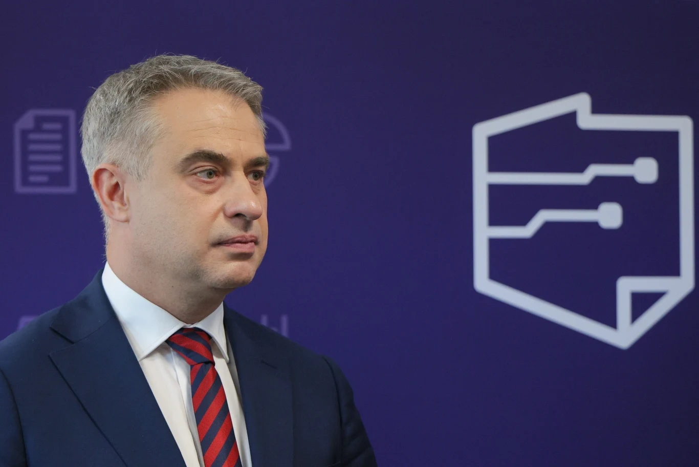 Krzysztof Gawkowski zapowiada start w wyborach na szefa Nowej Lewicy Mężczyzna w garniturze i czerwono-granatowym krawacie stoi na tle fioletowej ściany z graficznymi symbolami przedstawiającymi dokumenty i nowoczesne technologie.