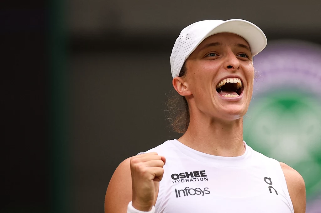 Iga Świątek Sportowiec w białym stroju i czapce z daszkiem, zaciśnięta pięść w geście triumfu, wyraz radości na twarzy, w tle rozmazane logo turnieju tenisowego
