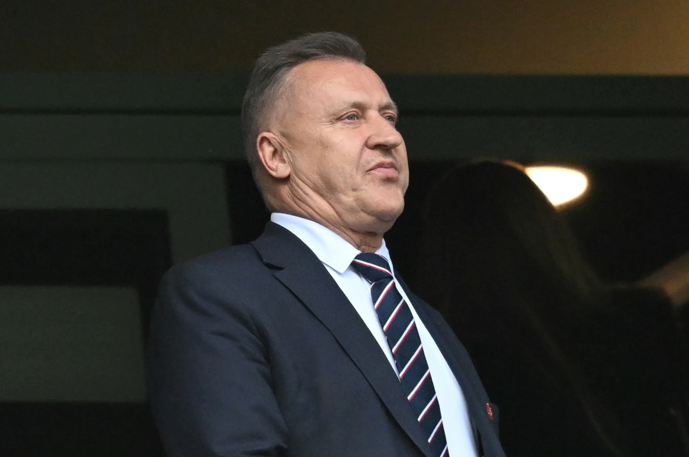 Mężczyzna w średnim wieku ubrany w garnitur i krawat w paski, stoi w pomieszczeniu z ciemnym tłem, w tle rozmazana postać i światło.