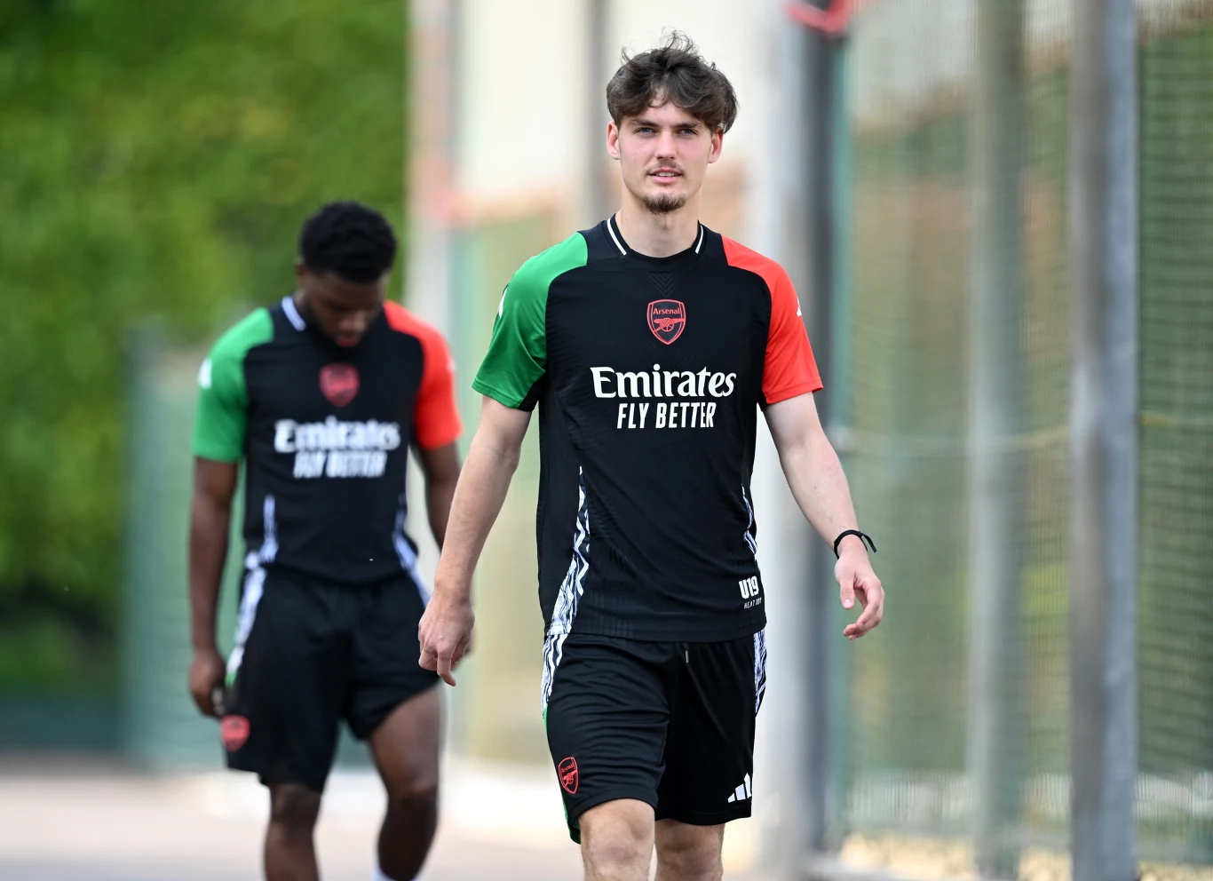 Michał Rosiak Dwóch młodych mężczyzn w sportowych strojach klubu Arsenal spaceruje obok boiska, obaj ubrani w czarne koszulki treningowe z logo klubu i sponsorów, jeden z nich idzie na pierwszym planie, drugi w tle.