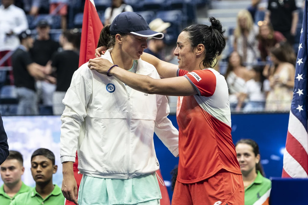 Iga Świątek i Ons Jabeur po finale US Open 20222 Dwie tenisistki po zakończonym meczu obejmują się na środku kortu, jedna z nich ubrana w białą kurtkę i czapkę z daszkiem, druga w czerwonym stroju sportowym, w tle sędziowie oraz publiczność.