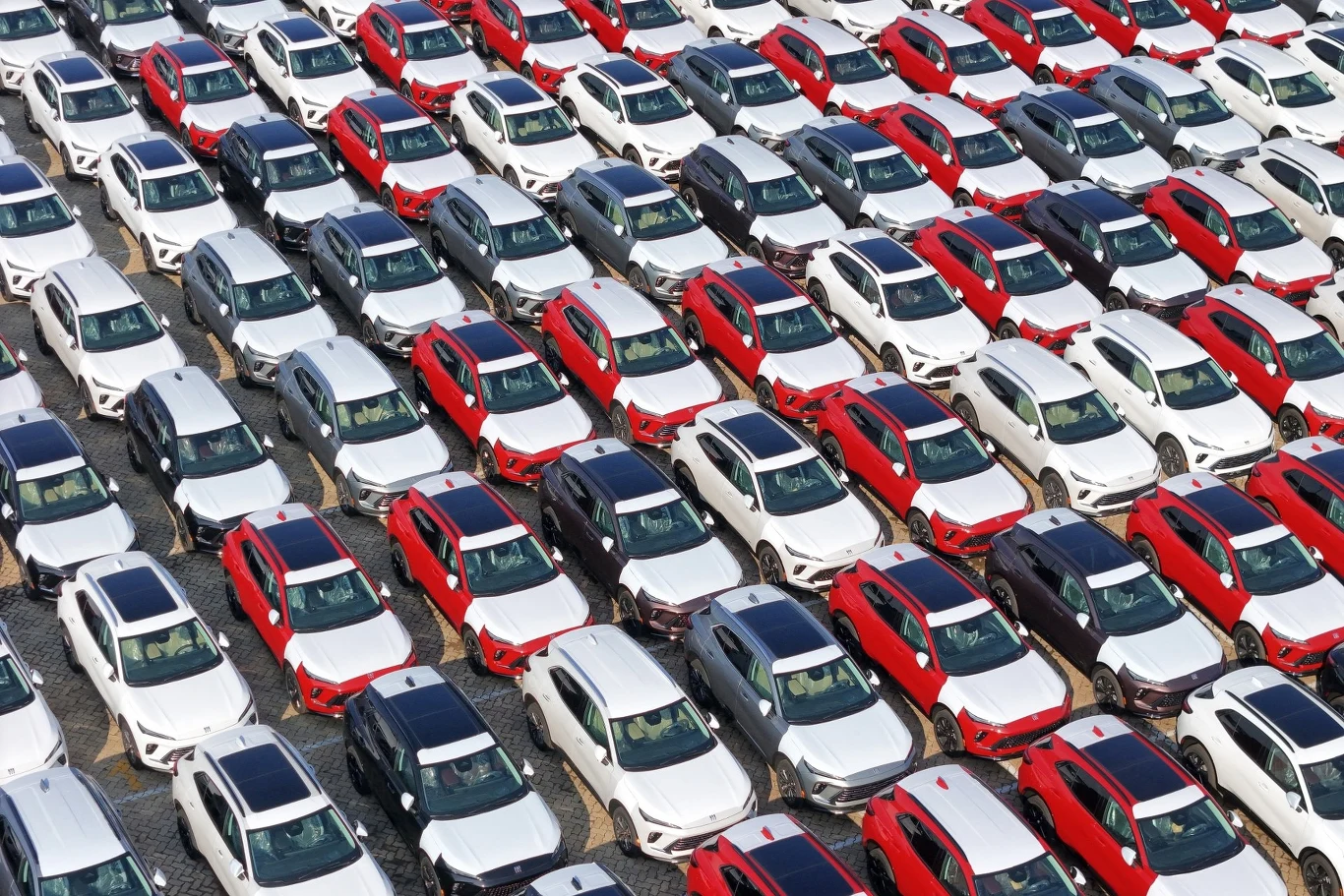 Chińska kontrola wykazała, że aż 21 725 pojazdów nie powinno było otrzymać dotacji. Szeregi nowych samochodów w kolorach białym i czerwonym ustawione równolegle na dużym placu, każdy pojazd zaparkowany bardzo blisko kolejnego, tworząc geometryczny wzór.