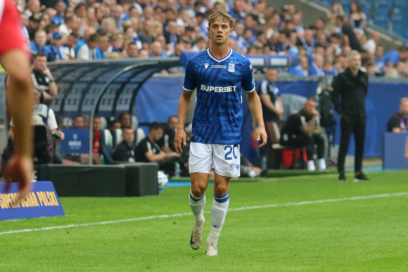 Piłkarz w niebieskiej koszulce z numerem 20 oraz napisem sponsorującym 'Superbet' stoi na murawie boiska podczas meczu piłki nożnej, w tle widoczna ławka rezerwowych oraz tłum kibiców na trybunach.