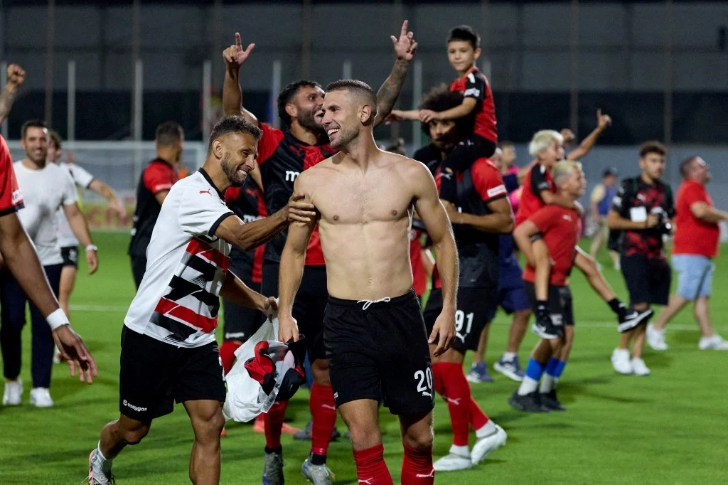 Hamrun Spartans FC świętuje awans do 2. rundy el. LM Piłkarze świętują zwycięstwo na boisku, jeden z nich stoi bez koszulki i uśmiecha się, a wokół zbiera się grupa zawodników i kibiców w klubowych barwach, wskazując rękami w górę z radości.