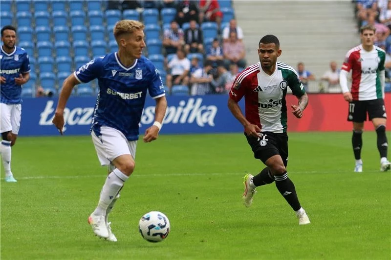 Lech Poznań - Legia Warszawa w meczu o Superpuchar Polski Piłkarze dwóch drużyn walczą o piłkę podczas meczu na stadionie, na trybunach widoczni kibice, a w tle banery reklamowe.