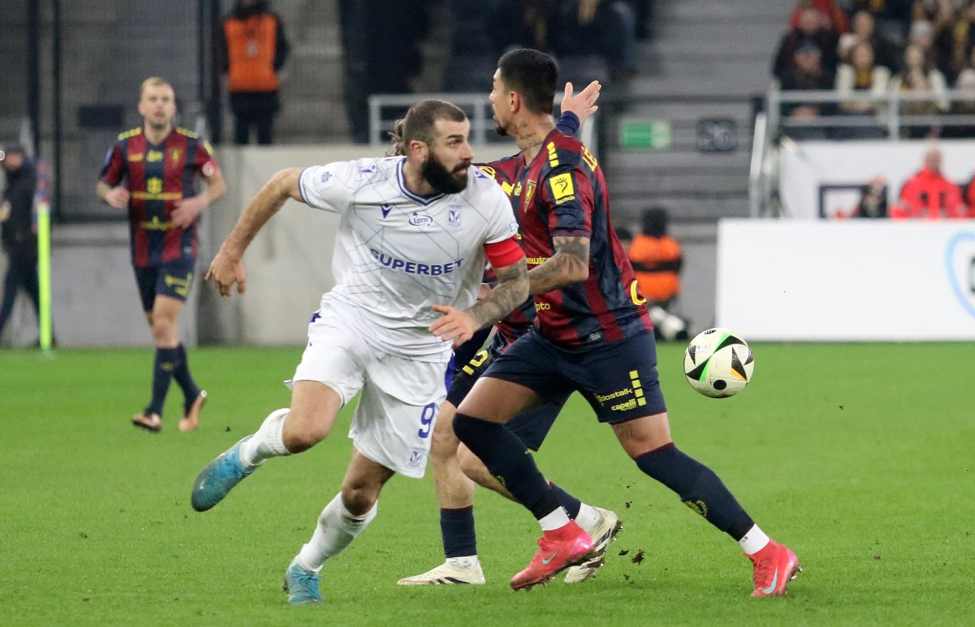 Lech Poznań - Pogoń Szczecin Dwóch piłkarzy w trakcie walki o piłkę na boisku, zawodnik w białym stroju ściera się fizycznie z rywalem w pasiastym czarno-granatowo-czerwonym kostiumie, w tle widoczny fragment stadionu oraz inni zawodnicy.