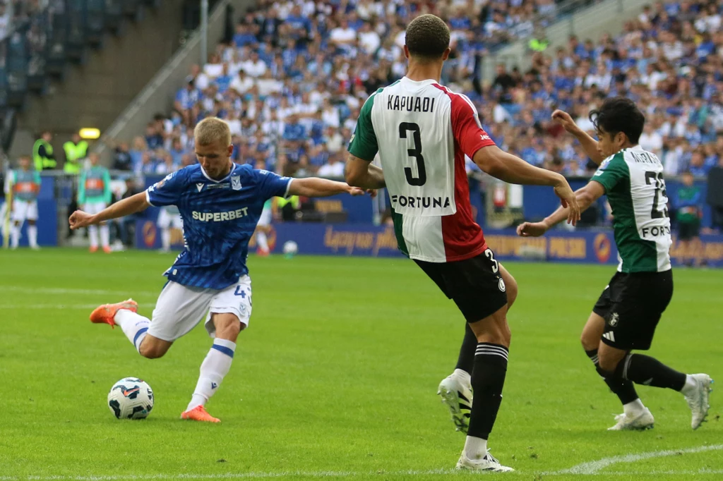 Lech Poznań - Legia Warszawa Zawodnik w niebieskiej koszulce strzela piłkę w kierunku bramki, a dwóch przeciwników próbuje zablokować akcję podczas meczu piłkarskiego na stadionie pełnym kibiców.