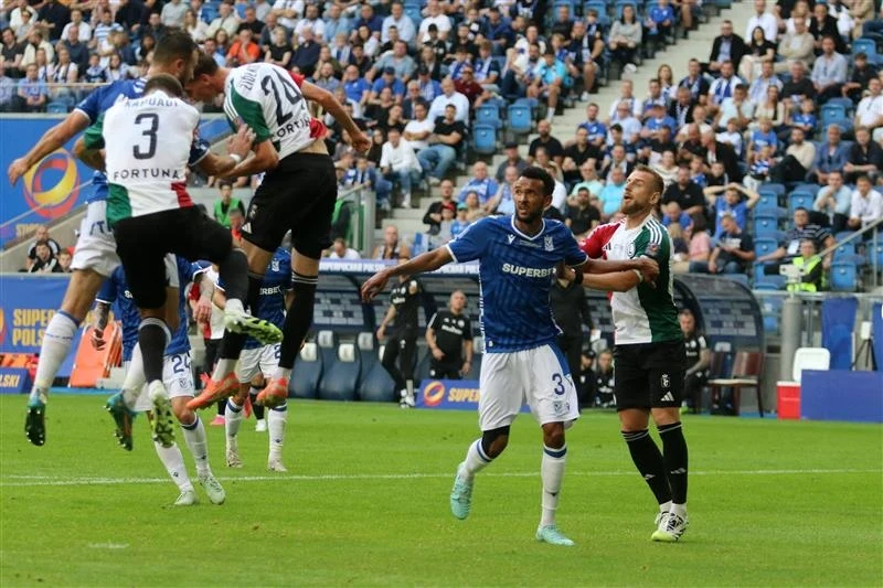 Lech Poznań - Legia Warszawa Piłkarze dwóch drużyn walczący o piłkę podczas rzutu rożnego na tle trybun wypełnionych kibicami, jeden zawodnik wyskakuje do główki, pozostali obserwują lub próbują przeszkadzać rywalowi.