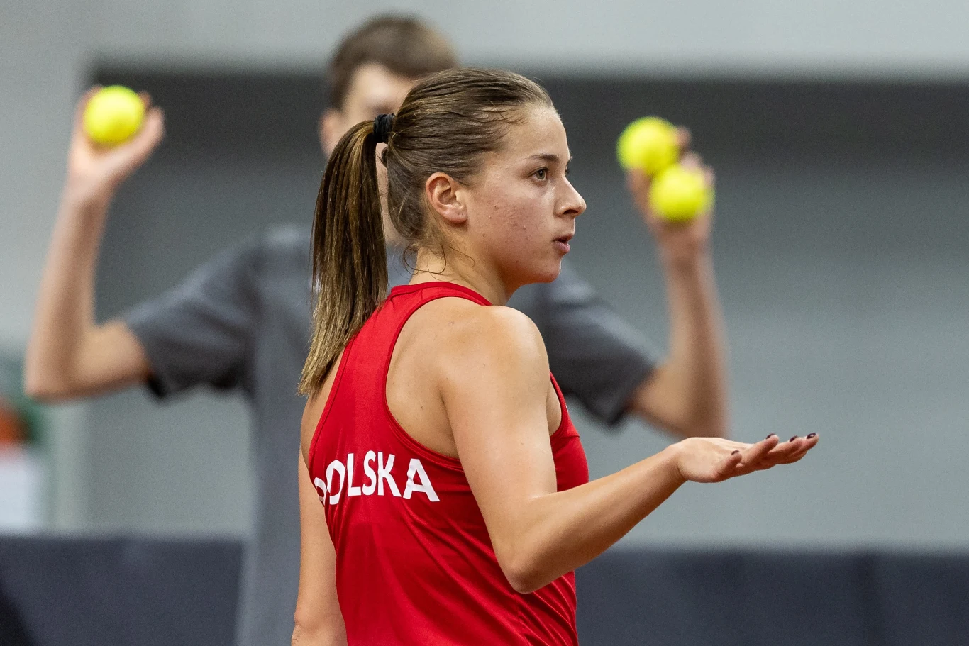 Kobieta w czerwonym stroju sportowym z napisem 'Polska' na plecach stoi na korcie, w tle osoba trzymająca trzy piłki tenisowe.