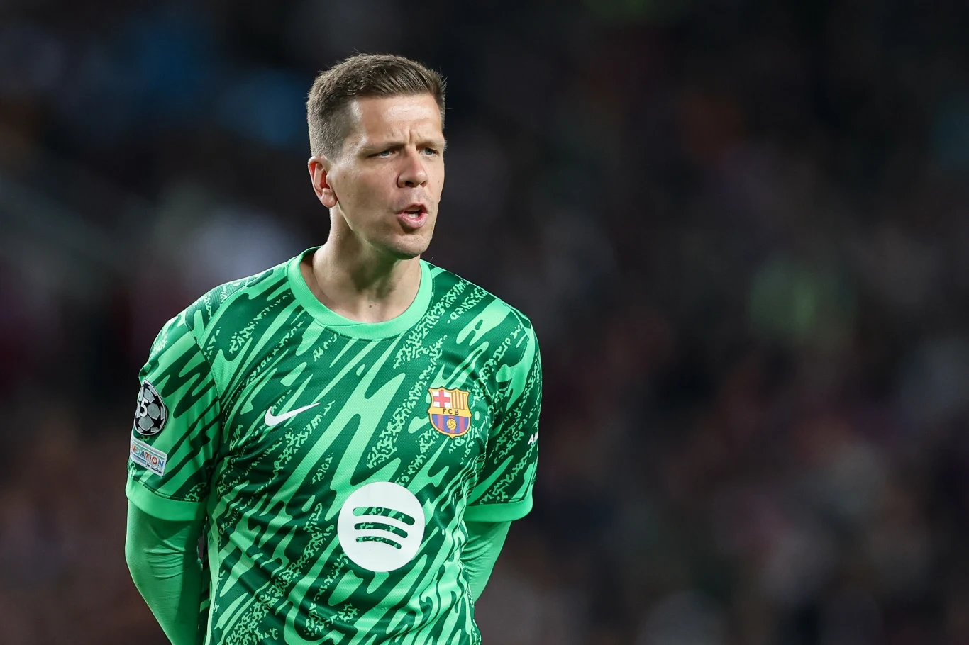 Wojciech Szczęsny Mężczyzna w zielonej koszulce bramkarskiej z herbem FC Barcelony oraz logo sponsora Spotify, stojący na tle rozmytego stadionu.