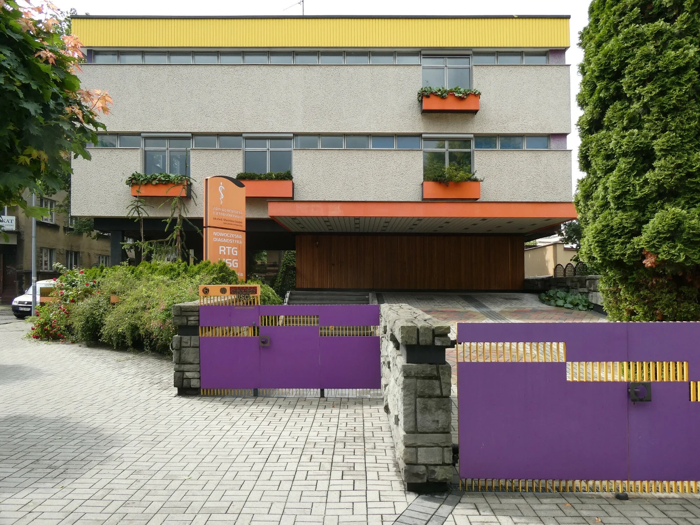 Nowoczesny budynek o prostych, geometrycznych kształtach, z szarymi ścianami i pomarańczowymi akcentami. Przed wejściem widoczna jest wysoka brama w kolorze fioletowym, a elewację zdobią doniczki z kwiatami. Po prawej stronie rośnie duże, zielone drzewo.