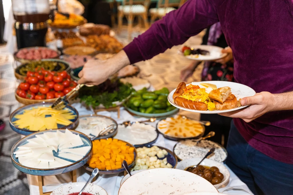 W hotelowych restauracjach lepiej uważać na surowe warzywa i owoce Ręka sięgająca po jedzenie z bogato zastawionego stołu, na którym znajdują się różne sery, pokrojone owoce, warzywa, ciasta oraz inne przystawki, a w tle widoczne są osoby oraz elementy sali.