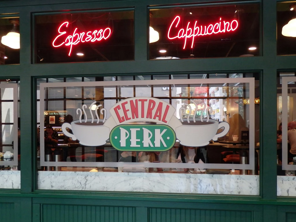 "Przyjaciele" Witryna kawiarni z dużym oknem ozdobionym napisem CENTRAL PERK oraz grafikami dwóch filiżanek z parą, powyżej świecące w neonach napisy Espresso i Cappuccino.