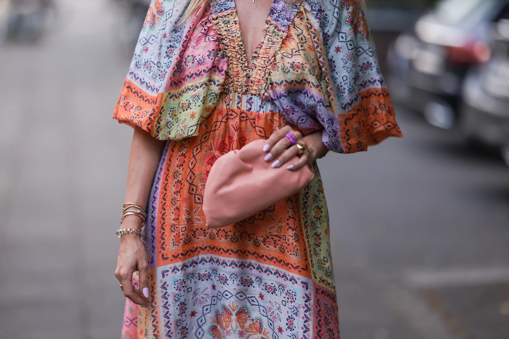 Sukienka typu kimono Kobieta w kolorowej sukience z etnicznym wzorem trzyma pastelową kopertówkę w jasnoróżowym kolorze, na jej dłoni widoczne są złote bransoletki oraz pierścionki, w tle rozmyta ulica i samochody.