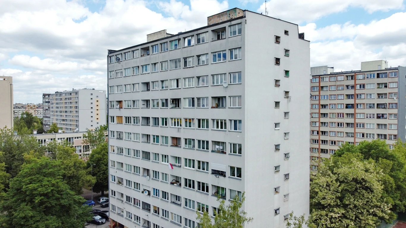Życie w wieżowcu: marzenie czy udręka? Wysoki blok mieszkalny z wielkiej płyty otoczony drzewami, w tle widoczne są inne podobne budynki mieszkalne oraz fragment osiedla z parkingiem.