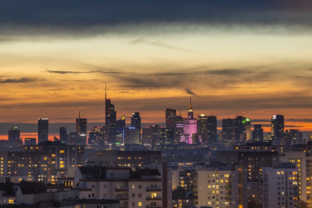 Panorama Warszawy z licznymi wieżowcami oświetlonymi światłami wieczornymi, charakterystyczny Pałac Kultury i Nauki podświetlony na fioletowo, ciemniejące niebo z pomarańczowo-niebieskim zachodem słońca.