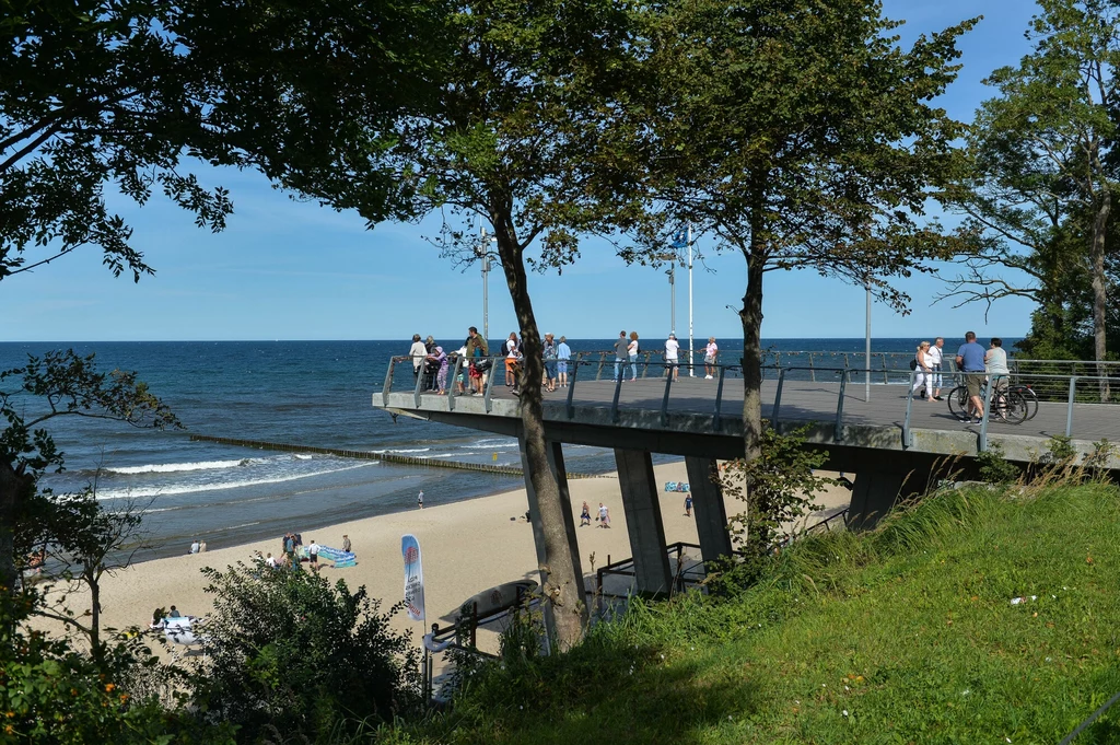 Molo wysunięte nad morze z grupą spacerujących ludzi, w tle piaszczysta plaża z wypoczywającymi osobami oraz spokojna, błękitna tafla wody. Wokół molo rosną drzewa, które tworzą lekki cień na trawie.