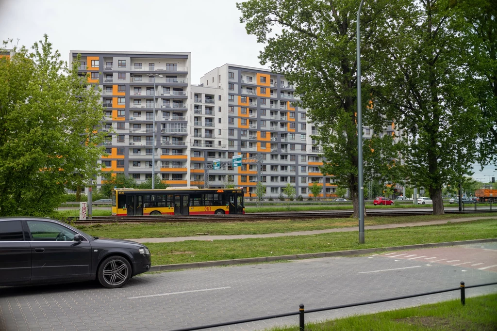 Nowoczesne osiedle mieszkaniowe z wysokimi blokami, charakterystycznymi pomarańczowymi akcentami na elewacjach, autobus komunikacji miejskiej jadący ulicą oraz osobowy samochód zaparkowany przy chodniku, otoczenie zielone przez drzewa i trawniki.