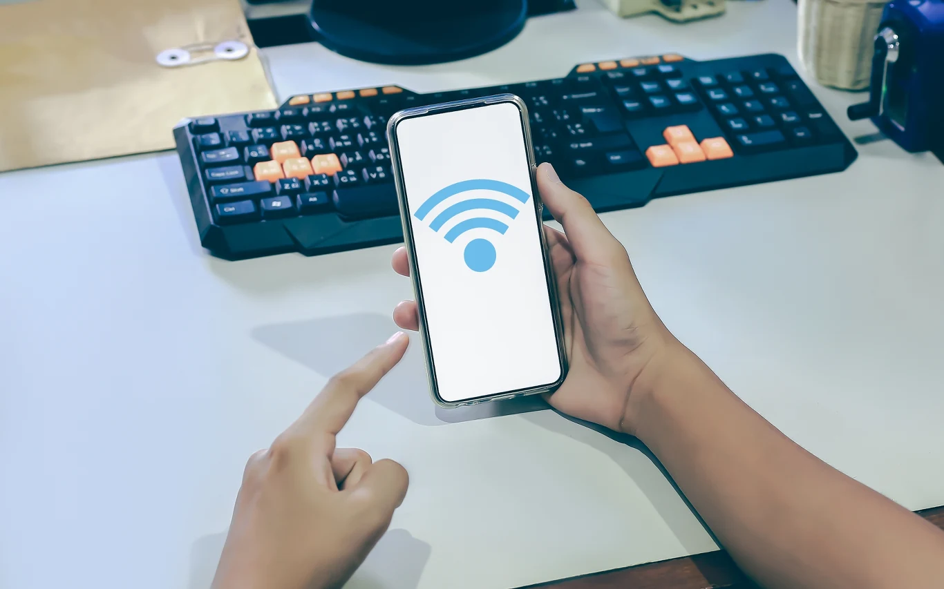 Wi-Fi słabo działa w mieszkaniu? W ten sposób pozbędziesz się czarnych dziur. Dłoń trzyma smartfon z wyświetlonym symbolem sieci Wi-Fi, palec wskazujący drugiej ręki skierowany w kierunku ekranu telefonu, w tle klawiatura komputerowa oraz monitor na biurku.