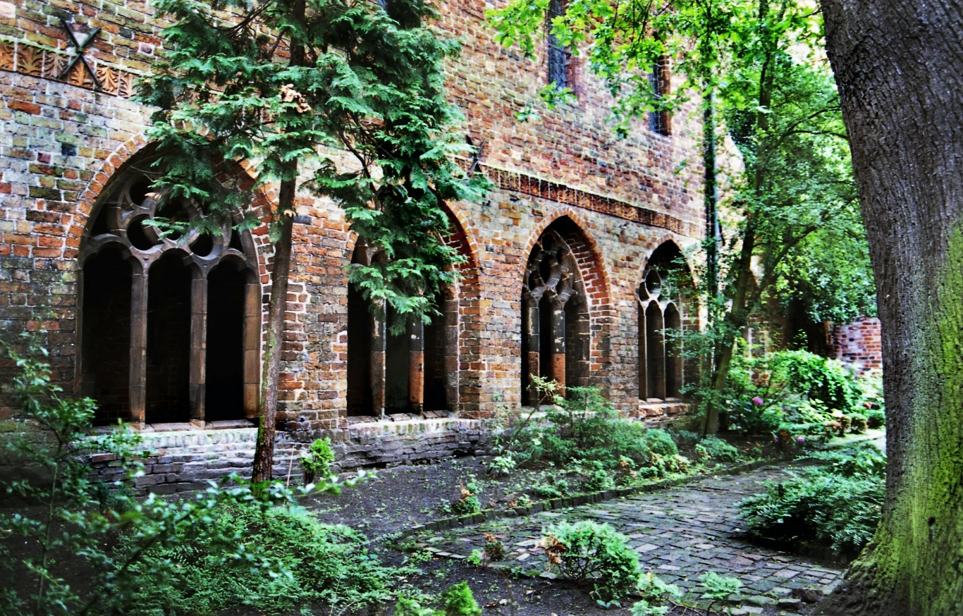 Gotyckie krużganki z ceglanymi arkadami otoczone bujną zielenią, stare drzewa i krzewy tworzą spokojny, historyczny klimat miejsca.