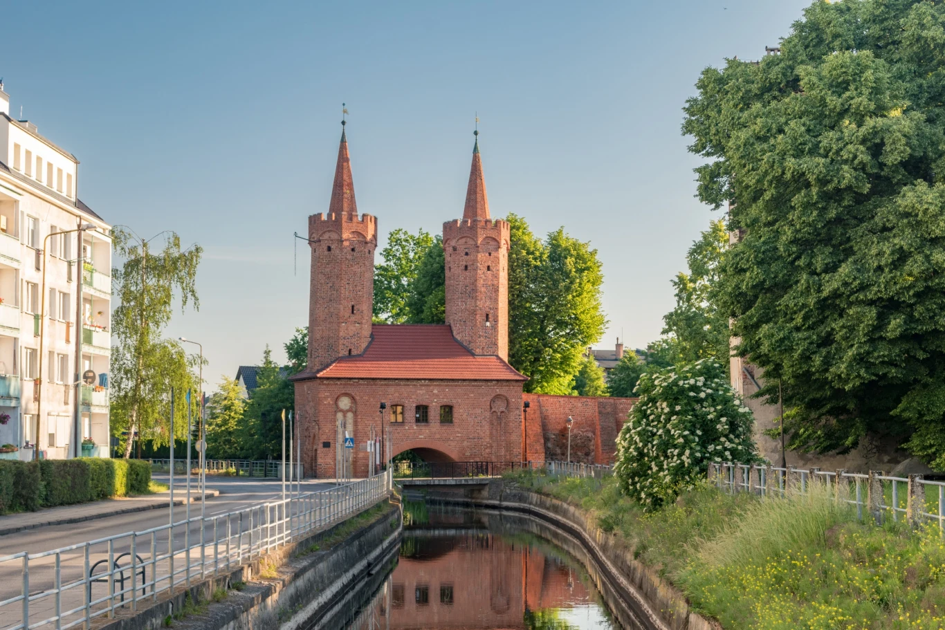 Gotycka brama miejska z dwiema wysokimi wieżami z czerwonej cegły, otoczona zielenią oraz wodnym kanałem, po bokach nowoczesne i historyczne budynki, spokojna atmosfera miejskiego krajobrazu.