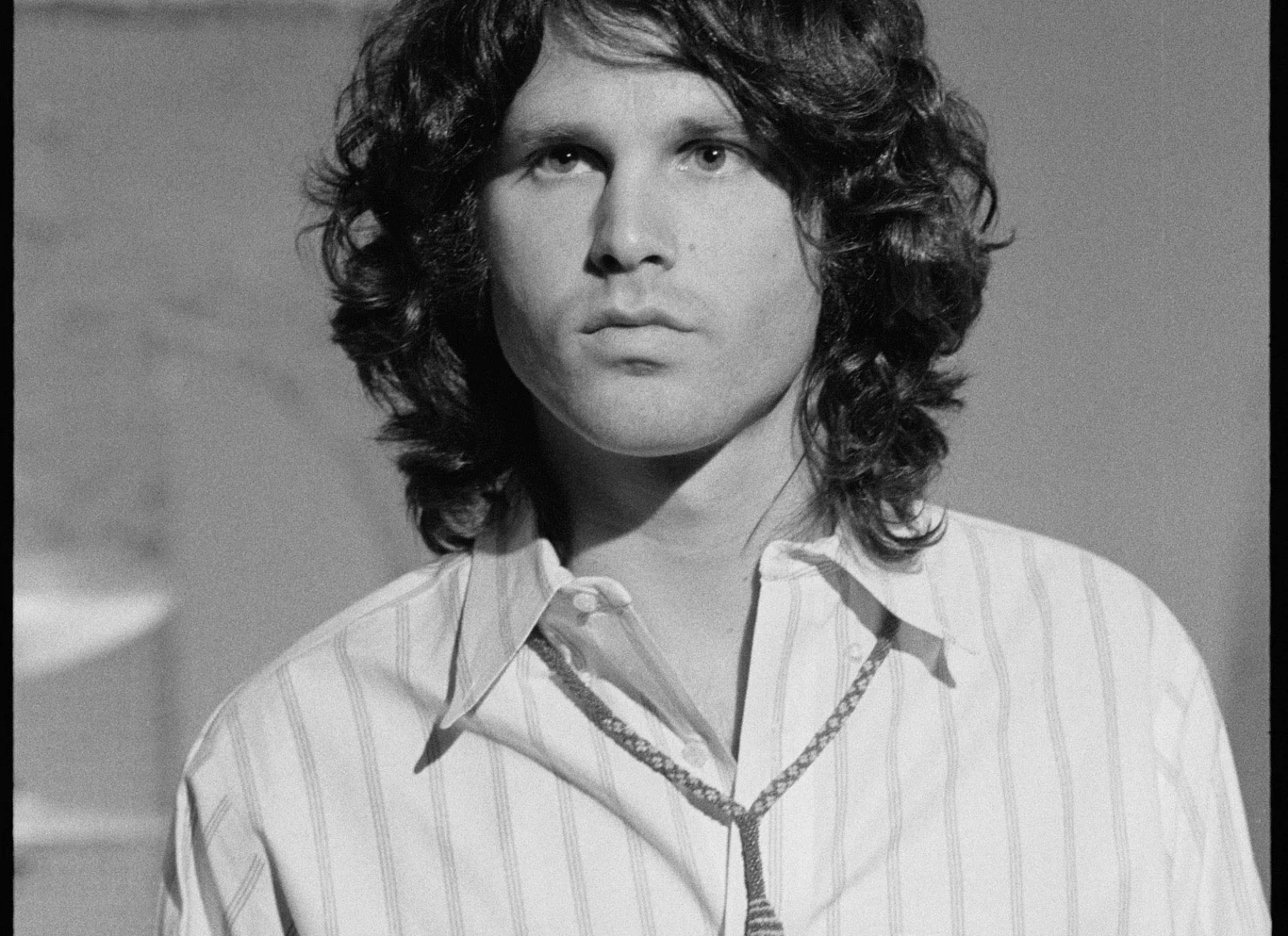 Jim Morrison (The Doors) - zdjęcie z 6 stycznia 1969 r. Mężczyzna z falowanymi, półdługimi włosami ubrany w jasną koszulę w paski, patrzący poważnie przed siebie, na szyi nosi naszyjnik z zawieszką.