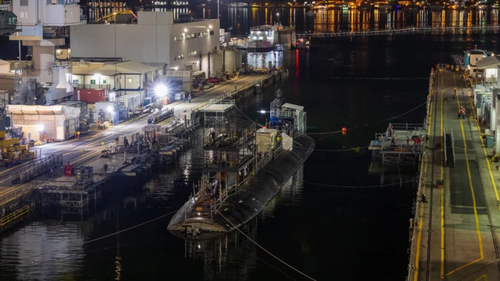 USS Connecticut zadokowany w Puget Sound Naval Shipyard, gdzie przechodzi naprawy, zdjęcie z grudnia 2023 roku. Można zauważyć brak kopuły dziobowej, która została zdjęta ze względu na uszkodzenia Duża łódź podwodna zacumowana przy nabrzeżu stoczniowym nad wodą, wokół niej rozstawione są rusztowania i maszyny, całość otoczona jest industrialną infrastrukturą oraz oświetleniem nocnym, w tle widoczne inne budynki i światła miasta.
