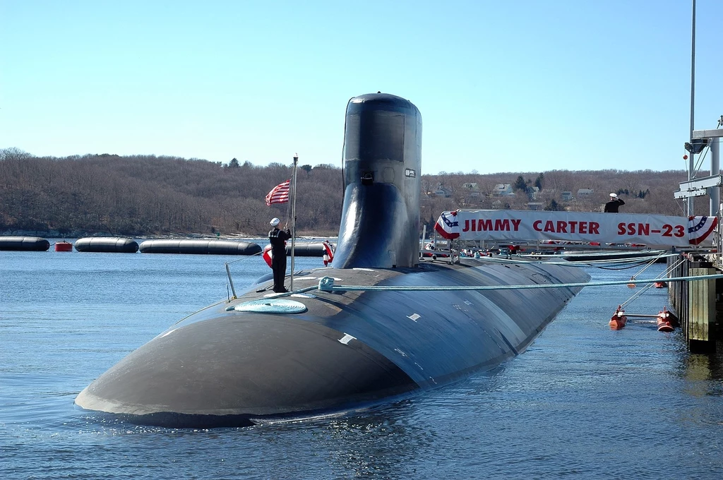 USS Jimmy Carter (SSN-23) Okręt podwodny klasy wojskowej cumujący przy nabrzeżu z flagą Stanów Zjednoczonych, obok baner z napisem Jimmy Carter SSN-23, w tle widoczne zalesione wzgórza