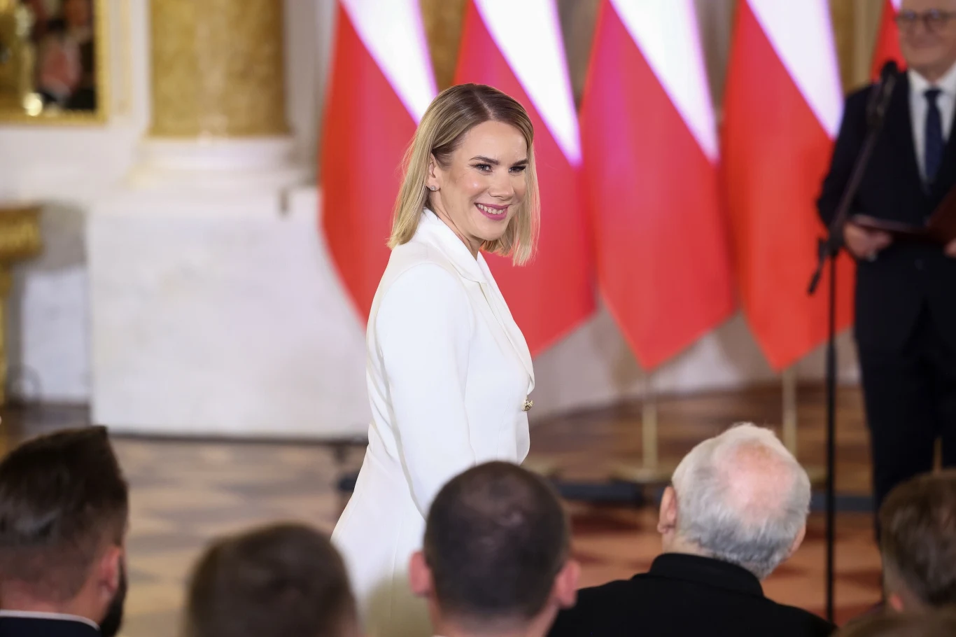 Kobieta w białym garniturze uśmiecha się do zgromadzonych osób na tle polskich flag podczas oficjalnego wydarzenia w eleganckiej sali.