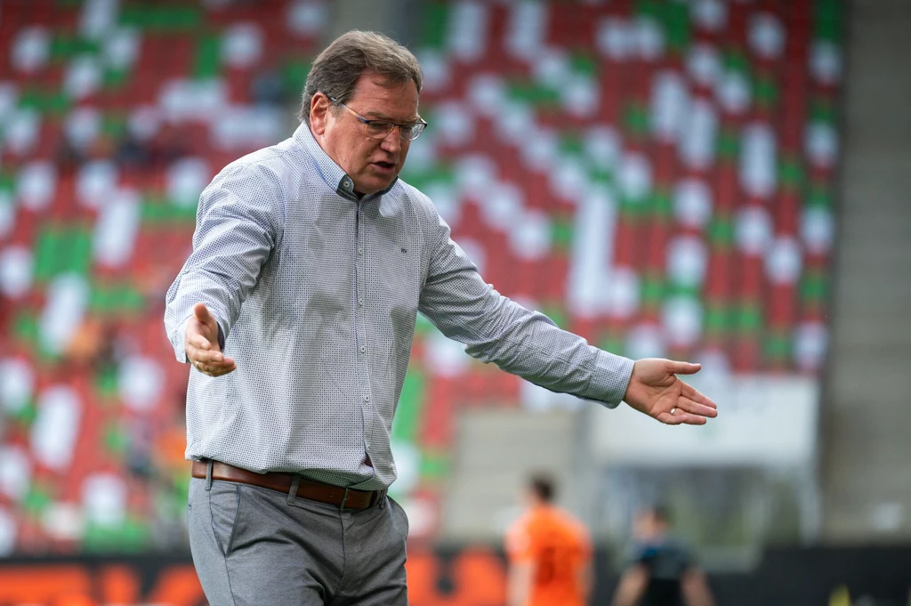 Jan Urban Mężczyzna w okularach, w jasnej koszuli i spodniach, gestykuluje z szeroko rozłożonymi rękami na tle stadionu z kolorowymi miejscami na trybunach.