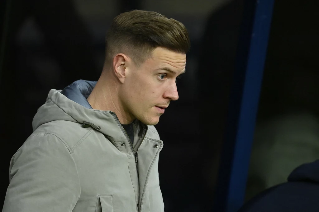 Marc-Andre ter Stegen Mężczyzna w szarej kurtce z krótkimi, starannie ułożonymi włosami patrzy w bok, stoi w pobliżu ciemnego tła.