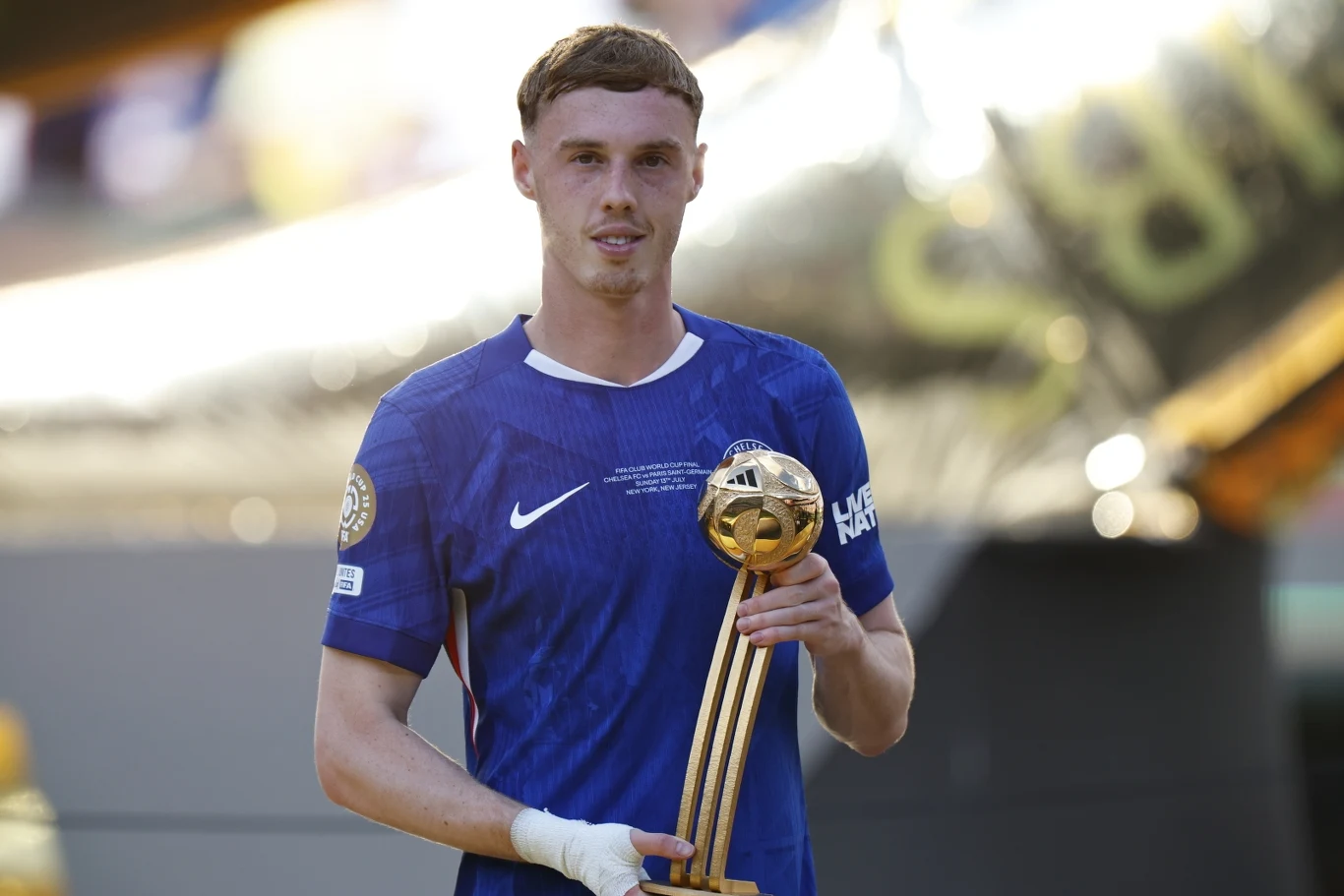 Młody mężczyzna w sportowej niebieskiej koszulce trzyma w rękach okazałe trofeum z kulistym złotym elementem, uśmiecha się lekko stojąc na stadionie w otoczeniu słonecznego światła.